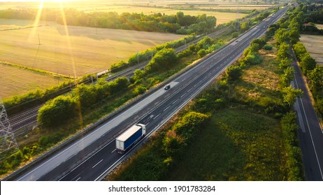 Aerial Drone Shot: Long Haul Semi Trucks Driving On The Busy Highway In The Rural Region Of Italy. Agricultural Crop Fields And Hills In The Background