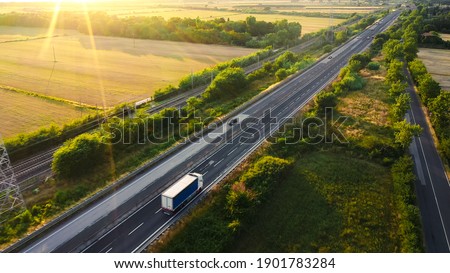 Aerial Drone Shot: Long Haul Semi Trucks Driving on the Busy Highway in the Rural Region of Italy. Agricultural Crop Fields and Hills in the Background