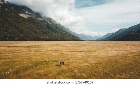 Aerial Drone Shot Of Two People Walking Amidst The Golden Grass In Eglinton Valley On The Way To Milford Sound In South Island New Zealand With Partly Cloudy Skies And Hills In The Background