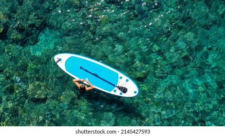 Aerial drone top down photo of fit woman woman relaxes in the sea next to a SUP board. Girl in sea with Stand Up Paddle board in tropical exotic turquoise clear sea.