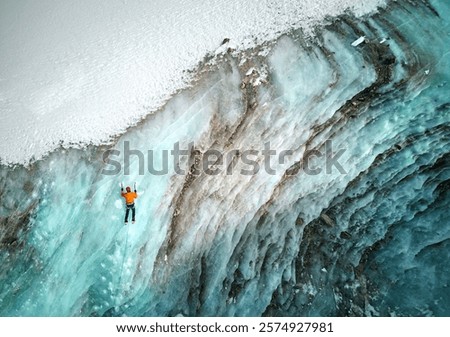 Aerial drone view of athlete in orange jacket Ice climbing at big frozen wall of ice ion Bogdanovich glacier in the mountain valley in Kazakhstan
