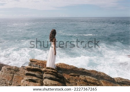 Aerial drone view of beautiful bride in an amazing white dress standing on a cliff and looking at the sea. Epic scene with stormy waves and gorgeous woman model