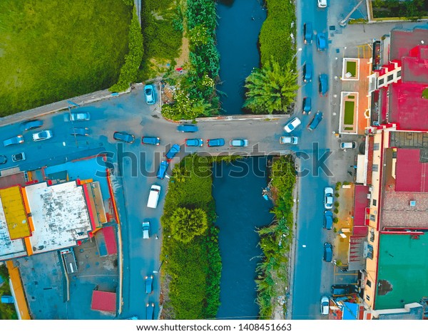 Aerial drone view of cars driving on a bridge\
crossing a river with colorful commercial buildings on either bank\
and a green field
