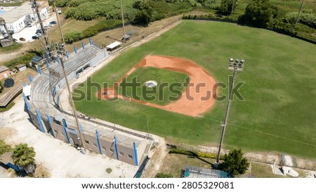 Aerial drone view of an empty baseball stadium on a sunny day, showcasing the distinctive red clay diamond, green grass outfield, concrete grandstands, pitcher's mound, and clear baseline markings.