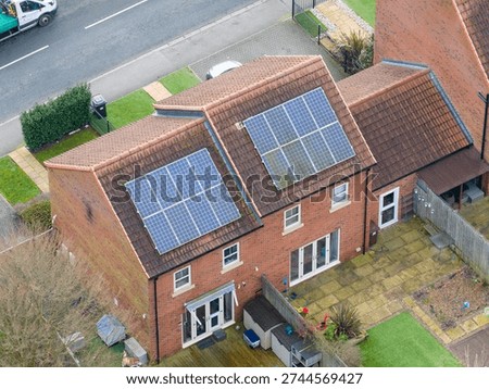 Aerial drone view looking down at modern brick suburban houses in the United Kingdom with rooftop solar panel systems producing renewable electricity for residential homes during winter season. The image shows sustainable living and green energy technology installed on tiled roofs of contemporary British housing development. One property features solar panels that appear dirty and partially covered with grime highlighting the need for seasonal maintenance cleaning and efficiency care for photovoltaic systems. Quiet neighbourhood streets small gardens patios and fenced backyards surround the homes creating a typical UK residential environment. The photograph represents clean energy adoption climate conscious living home energy independence and the everyday reality of solar technology installed on family housing while also illustrating how weather dirt and winter conditions can affect panel performance across modern sustainable communities.