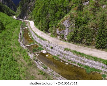 Aerial drone view of Lotru river flowing through a narrow canal, narrowed by dams in a mountainous forested area. A gravel road winds along the river. Sharp cliffs lay the valley. Carpathia, Romania. 