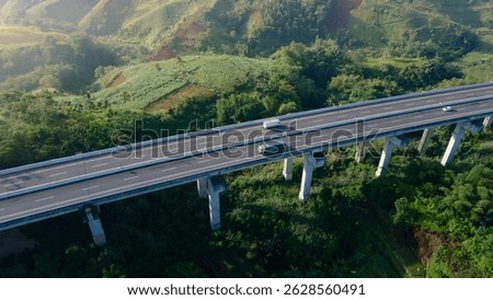 Aerial drone view of a modern elevated highway bridge crossing over scenic green hills. Several vehicles, including trucks and cars, are seen traversing the structure