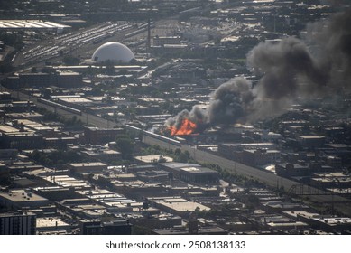 aerial elevated view of a massive building on fire, engulfed in flames in a dense urban environment with smoke billowing into the air