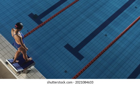 Aerial: Female Swimmer Stands on a Starting Block, ready to Jumps into Swimming Pool. Professional Athlete Swims, Determined to Win Championship. Dark Dramatic Light, High Angle View.