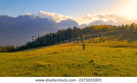 AERIAL: Fit young lady biking towards picturesque mountaintop with her brown dog. She is riding an electric bike along scenic mountain landscape which is glowing in autumn colors at golden sunset.