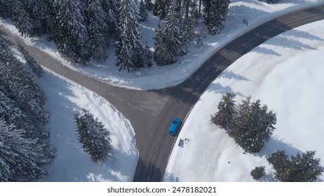 AERIAL: Flying above turquoise car driving along snowy road leading through dense evergreen mountain forest in winter. Road trip in picturesque pine tree woodland covered with fresh snow on sunny day - Powered by Shutterstock