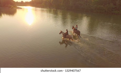 AERIAL: Flying Above Two Active Women Riding Their Horses Down The Tranquil River On A Picturesque Fall Morning. Carefree Girls Having Fun During A Relaxing Horseback Ride In The Serene Autumn Nature.