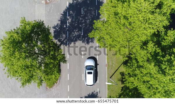 Aerial following car top-down view this grey\
colored station wagon is driving over two way street with green\
trees on both sides of\
street