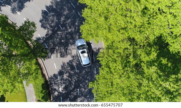 Aerial following car top-down view this grey\
colored station wagon is driving over two way street corner green\
trees on both sides of\
street