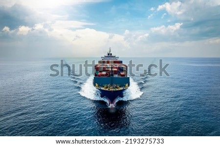 Aerial front view of a medium sized, loaded container cargo vessel traveling with speed over the ocean under cloudy sky