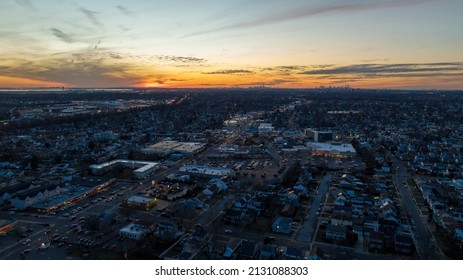 Aerial, High Angle Drone View High Over A Busy Long Island Town During A Colorful Sunset. Street Lights Are Turned On. The Roads Have Some Traffic And Movement.