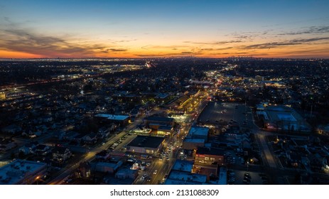 Aerial, High Angle Drone View High Over A Busy Long Island Town During A Colorful Sunset. Street Lights Are Turned On. The Roads Have Some Traffic And Movement.