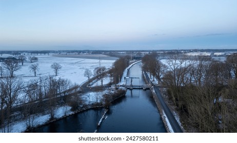 The aerial image captures the serene flow of a canal as it cuts through a vast expanse of snow-covered fields at dusk. The canal, flanked by roads and rows of bare trees, acts as a lifeline in the - Powered by Shutterstock