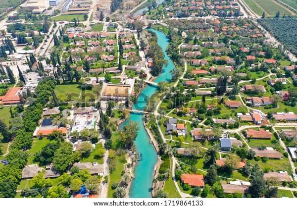 Aerial image of Kibbutz Nir David with Asi river\
channel turquoise water dividing east and west side riverside\
houses and palm trees,\
Israel.