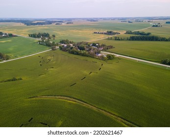 Aerial Landscape View Of Farm Fields. Country Road Intersection With Groups Of Trees And Buildings. Designs In The Field With A Eye. Hazy Blue Sky Overhead. More Buildings Scattered Through The Scene.