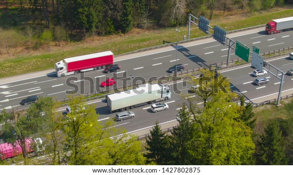 AERIAL: Large white cargo truck carrying a\
shipping container gets stuck in traffic on the busy freeway.\
Countless cars surround the big lorry transporting a heavy freight\
container to its\
destination