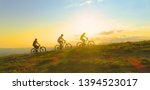 AERIAL LENS FLARE COPY SPACE SILHOUETTE: Fit tourists riding electric bicycles along a grassy path on a beautiful sunny spring day. Cinematic shot of three friends enjoying a scenic mountain bike ride