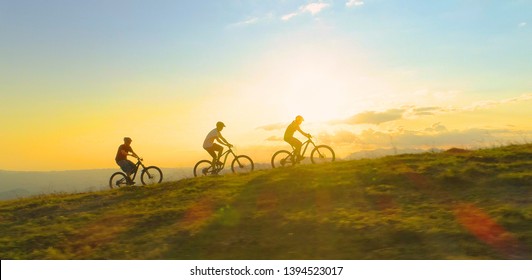AERIAL LENS FLARE COPY SPACE SILHOUETTE: Fit tourists riding electric bicycles along a grassy path on a beautiful sunny spring day. Cinematic shot of three friends enjoying a scenic mountain bike ride