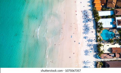 Aerial Overhead View Of Green Foaming Waves Crushing Against The Sandy Coast Line, People Enjoy Summer  Near Resorts And Hotel Line