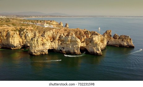 Aerial panoramic view of yachts sailing around white cliffs on ocean water surface with city placed on top of hills in sunset time, Portugal, Algarve.