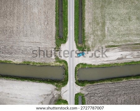 An aerial perspective captures a rural intersection marked by a blue pickup truck positioned at the crossroads of two gravel roads. Surrounding the intersection are fields with varying shades of brown and green, indicating recent agricultural activity. Two narrow water canals run alongside, reflecting the sunlight. The lushness of the farmland suggests it is late spring, a time of growth and renewal.