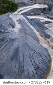 Aerial perspective of a large mining operation featuring dark mineral piles, textured grey soil, and sandy industrial roads with tire patterns from site vehicles.
