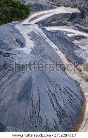 Aerial perspective of a large mining operation featuring dark mineral piles, textured grey soil, and sandy industrial roads with tire patterns from site vehicles.