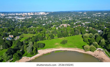 An aerial perspective recedes from two boys playing soccer in an open field within a picturesque park on a sunny day, transitioning to a wide-ranging view of the surrounding area with the city skyline - Powered by Shutterstock