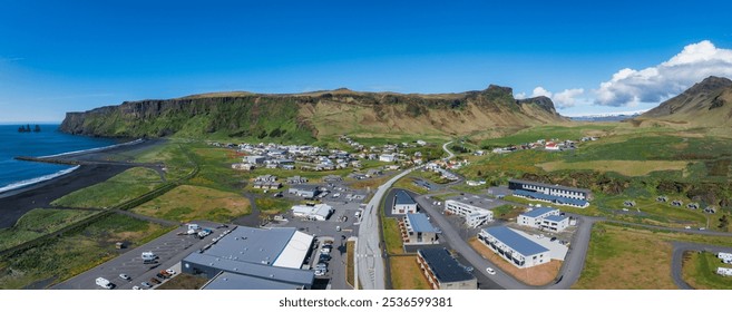 Aerial perspective of Vik i Myrdal, Iceland, featuring black sand beach, Reynisdrangar sea stacks, green cliffs, and a mix of buildings under a blue sky. - Powered by Shutterstock