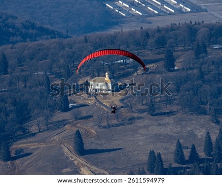 An aerial photo captures a paraglider with a red canopy flying above a winter landscape of bare trees and a prominent building with a dome. The structures add a human element to the natural setting.