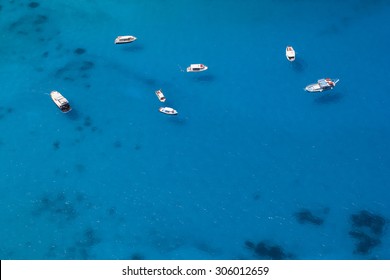 Aerial photo of several boats anchored in a sea bay with shallow clear blue water. The water is clean enough to enable to see the boats shadows on the seabed. 