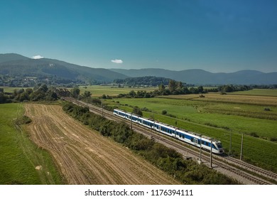 Aerial photo of white and blue passenger train running between Udine, Trieste and Ljubljana close to Preserje train station driving over a small bridge on a sunny day. - Shutterstock ID 1176394111
