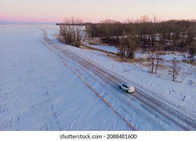 Aerial Photo Of A White SUV Driving Down A Snowy Road In Rural North Dakota. Snow Is Blowing Across The Road And Barren, Brown Vegetation Surrounds The Road. Beautiful Sunset. Winter.