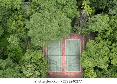 Aerial photograph captures  red clay tennis court nestled in the midst of a dense forest. The court is marked with white lines and surrounded by tall, lush trees that provide shade for the players. - Powered by Shutterstock