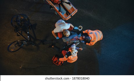 Aerial Portrait of a Young Injured Man Involved in Bicycle Accident is Being Saved by Medical Team of EMS Paramedics on the Street at Night. Emergency Care Assistants Provide Essential First Aid Help. Blur