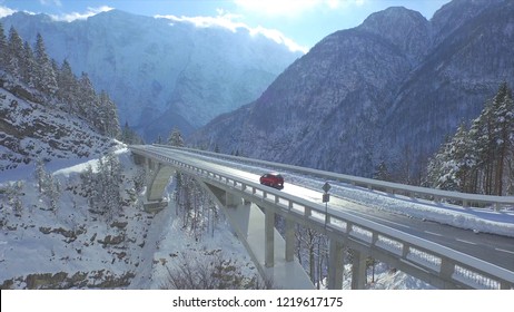 AERIAL: Red Car Crosses The Concrete Bridge Build In The Idyllic Snowy Wilderness. Flying Behind Tourists On A Fun Road Trip Driving Their Car Through The Spectacular Snow Covered Nature In Slovenia.
