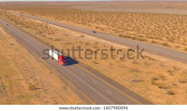 AERIAL: Red cargo truck hauls a heavy\
container across the barren landscape. Aerial shot of a\
semi-trailer truck transporting merchandise across the rugged Utah\
desert on a sunny spring\
afternoon.
