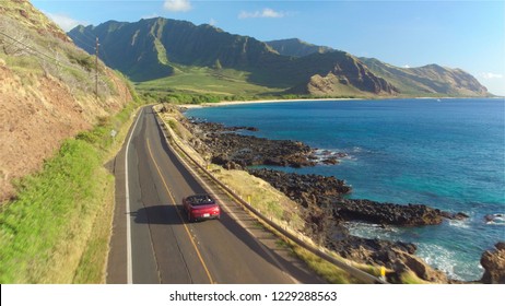AERIAL: Red convertible car driving along the picturesque coastal road above dramatic rocky shore towards beautiful volcanic mountains. Happy young couple on summer vacation traveling at the seaside