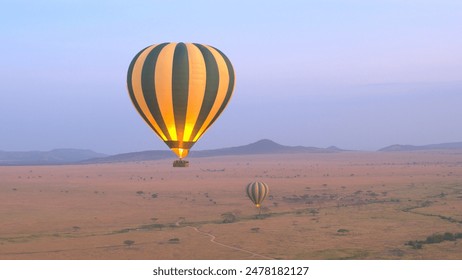 AERIAL: Safari hot air balloon flying above vast savanna plain rolling into the distance in Serengeti National Park at purple light dawn. Tourists traveling in air over African wilderness at sunrise - Powered by Shutterstock