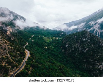 Aerial Scenery View Of Asphalt Road Pass In Rocky Mountains With Green Vegetation Covered With Fog After Rain. Bird's Eye View Of Rocky Hills With Picturesque Bike Path Above Which Hung Heavy Clouds