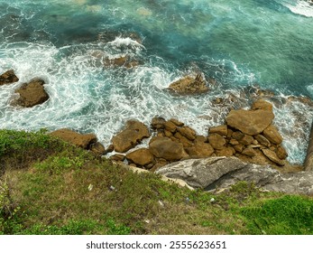 An aerial sea landscape view of a rocky coastline with vibrant turquoise waves crashing against the shore, surrounded by lush green grass - Powered by Shutterstock