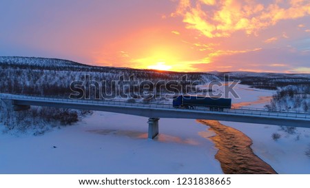 AERIAL: Semi truck crossing the bridge above icy river in the winter at sunset