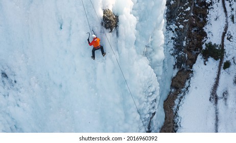 Aerial Shot Of A Frozen Waterfall, And A Caucasian Ice Climber Facing The Slope And Kicking His Toes In To Plant The Crampoins