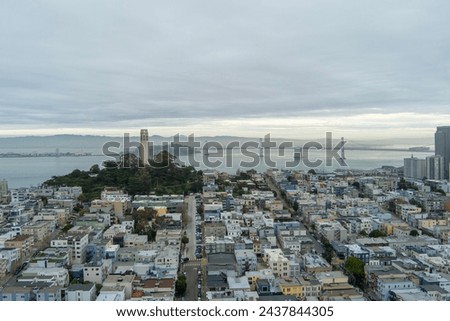 aerial shot of the ocean and streets lined with apartments, lush green trees and Coit Tower, the bay bridge over the ocean with powerful clouds at sunset in San Francisco California USA