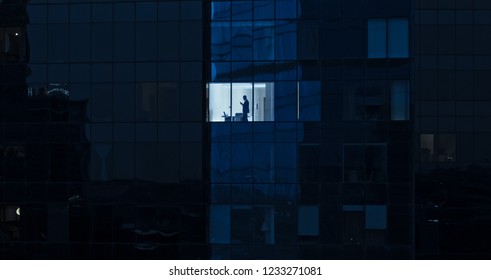 Aerial Shot: From Outside Into Office Building With Businessman Using Mobile Phone And Standing By The Office Window. Beautiful Shot Of The Financial Business District Skyscrapers.